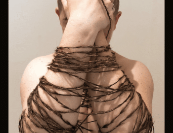 A person with a shaved head is wrapped in copper wire and their own hair, with arms crossed over their face, hiding their eyes. Text at the bottom describes the artwork as "Buzz Buzz: Beauty/Bound" by Liz Ashe.